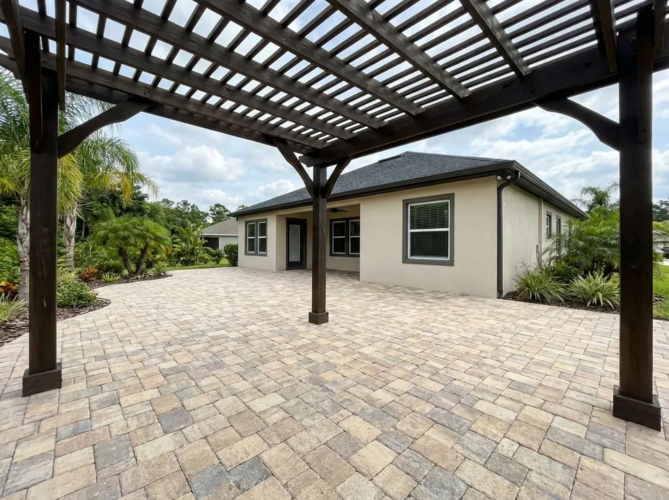 Tan tumbled-paver patio shaded by a dark-stained pergola off the rear of a one-story home