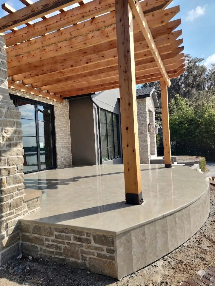 Curved raised terrace faced in stacked-stone veneer, paved in large-format pavers, under a wood pergola at the water
